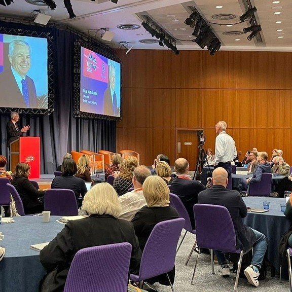 BTA Chairman Nick de Bois CBE stands behind a podium onstage and speaks to a room of 80 delegates at the VisitBritain Business Events Association Conference