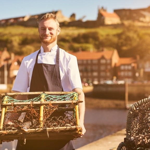 Chef wearing apron on pier holding lobster trap with lobster