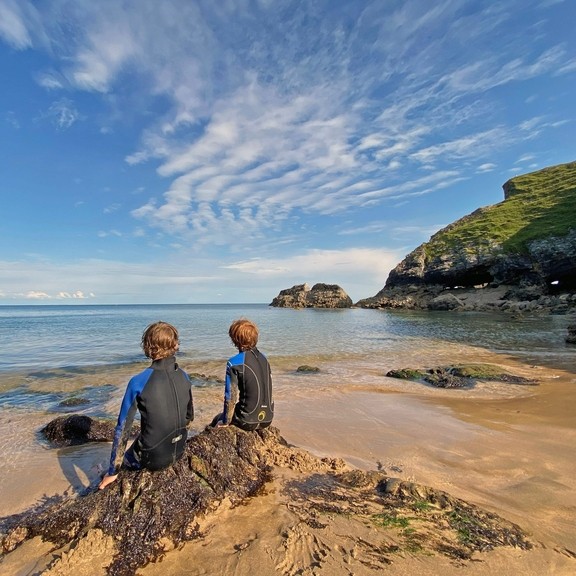 Back view of two young boys in wetsuits sitting on a rock looking out to sea with blue sky