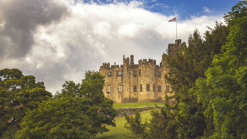 A large castle with a flag at full mast as seen through the gardens and trees.