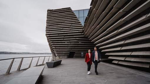 A man and a woman walking at the back of a museum with a sea view.