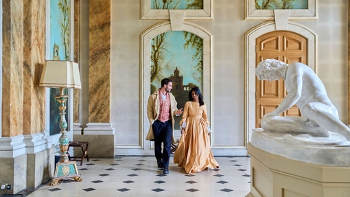 A man and a woman walk through a gallery in costumes at a heritage building