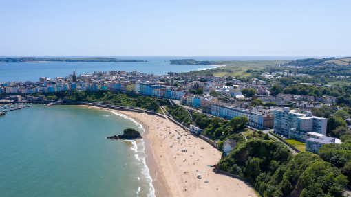 An overhead shot of a sandy beach lined with sunbathers, surrounded by brightly coloured townhouses and countryside.
