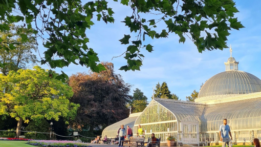 Large domed glass house building in the background in the sunshine, with people outside walking though the park, flower beds in foreground.