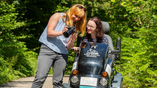 A woman stands holding a camera showing the pictures to a woman who sits in a tramper