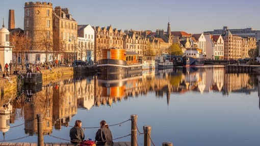 Two young women sat on the jetty of the waterfront in the evening with boats and buildings by the harbour.