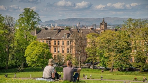 A couple sitting on a hill in park, with Glasgow skyline, and people cycling and walking along park paths.