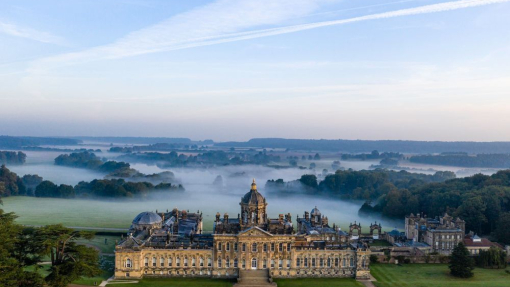 Large country house seen from above with formal gardens and wooded countryside behind covered in low lying cloud