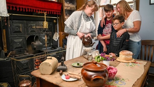 A group observing a kitchen scene from the past with a lady pouring tea at the table inside a living museum.