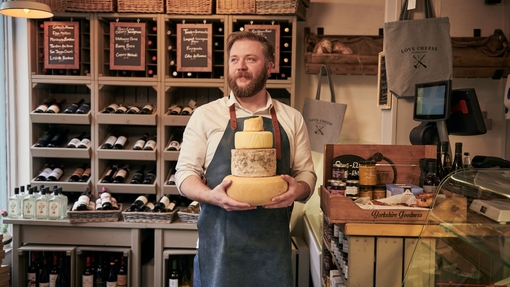 Man wearing blue apron holding a stack of cheeses in a cheese and wine shop