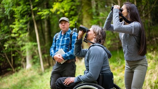 Man in checked shirt with telescope is telling two people, a woman using binoculars and a man using a wheelchair about the forest bird song at Dalby Forest.