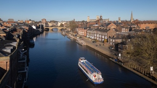 A boat travels down a river towards a bridge with the City beyond