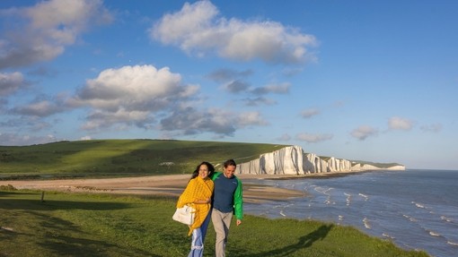 A couple walk together on a headland with white chalk cliffs in the background.