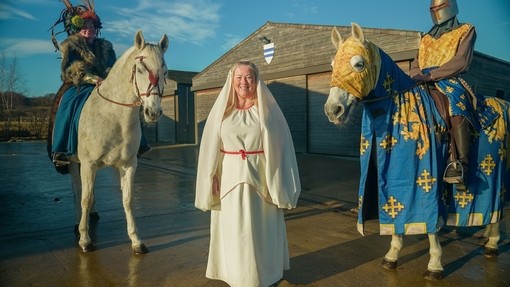 A woman poses with two people horseback all wearing medieval costume