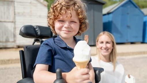 Young electric wheelchair user holding an ice cream cone with mum in background at the seaside