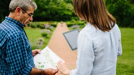 Rear view of male gardener sharing garden plans with female garden designer holding an iPad stood on a path in a garden