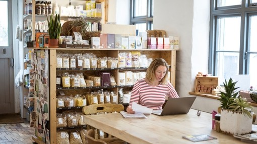 Caucasian businesswoman in early 40s using laptop to sort out paperwork while sitting at communal table in her health food shop and cafe
