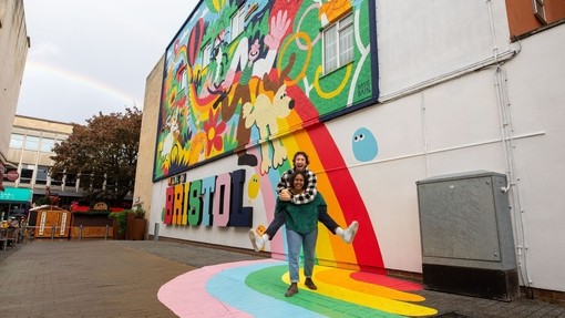 Two people playfully smiling and posing in front of a colourful mural.