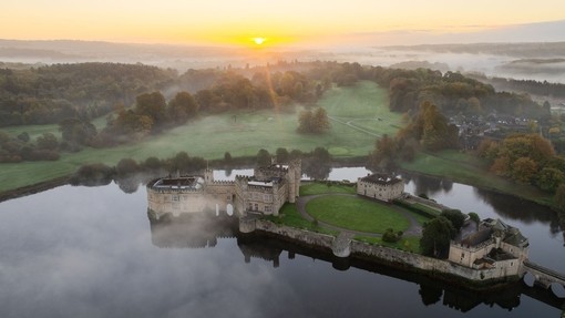 Aerial view of castle surrounded by a moat, river and trees, enveloped in mist during sunrise