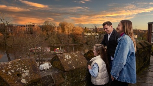 A family of three look out over a town river from a bridge.