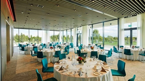 A number of round dining tables are laid with white table clothes and set for a meal. The room has floor to ceiling windows which look out on fields and trees. National Memorial Arboretum - Gold award winner for the Business Events Venue of the Year at the VisitEngland Awards for Excellence 2023.