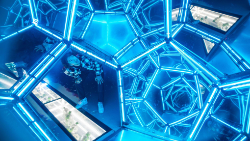 Man looking up at his reflection inside tunnel with hexagonal patterned walls and blue lighting