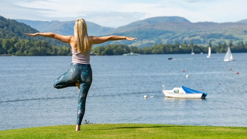 A woman stands in a yoga pose at the side of a lake with mountains beyond