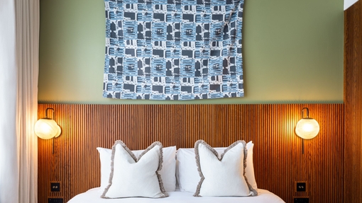 A hotel room bed with crisp white sheets and pillows with a wooden headboard against a green wall