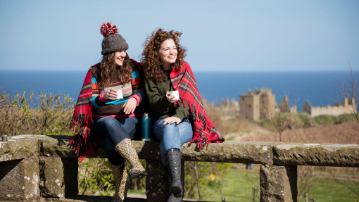 Two women with hot drinks, sitting on a stone wall near a castle