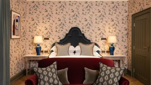 Plush hotel bedroom with red velvet sofa at the foot of a bed with flowery wallpaper on the walls behind it.