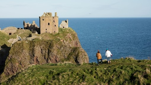 Two women walking along a grassy clifftop, near a castle