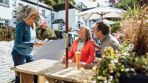 A couple sitting at a wooden table in a beer garden, while a waitress wearing a Covid mask takes their order