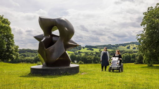 Two women look at a large sculpture set in green gardens with landscape beyond