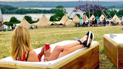 Young blond woman lying in a bath overlooking festival tents, with hills and water in the distance