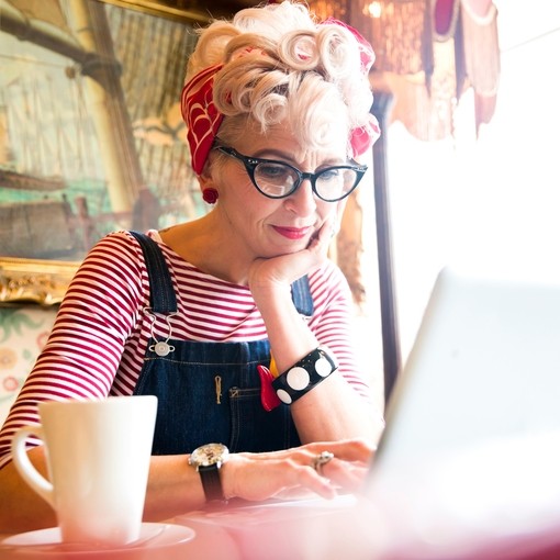 Close up of a blond woman wearing a stripy top and red head scarf, having a coffee and looking down at a lap top screen, smiling.