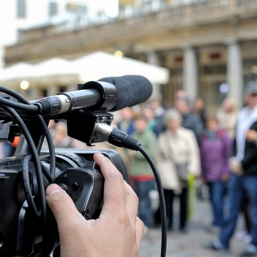 Close up image from the rear of a TV camera with an external directional microphone and XLR lead mounted on the camera. The camera operators hand on the focus hand grip with out of focus people in the background.