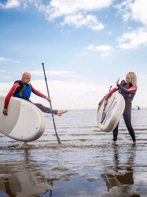 A man and woman in wet suits stand holding paddleboards on a beach