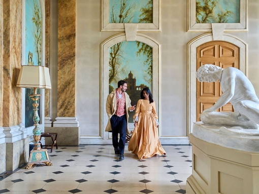 A man and a woman walk through a gallery in costumes at a heritage building