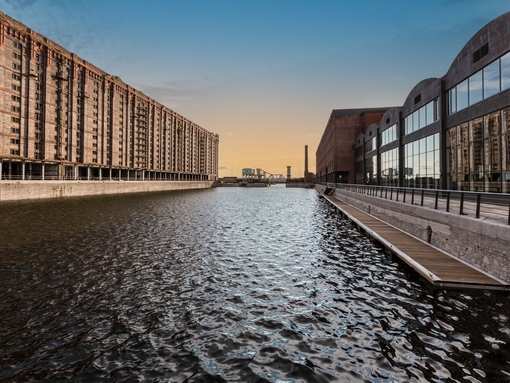 A view of a dock looking across the water. To the left stands a traditional brick built dockside building, to the right is a modern building made from glass and wood. 