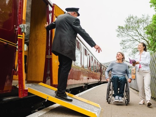 Guard stands on a ramps and leans out of heritage train carriage greeting woman and man using a wheelchair
