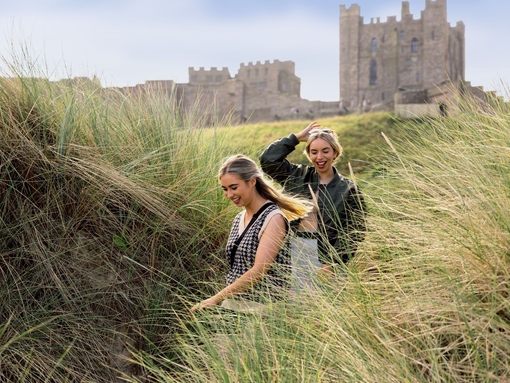 Two women walking in the sand dunes by the sea with a castle in the background