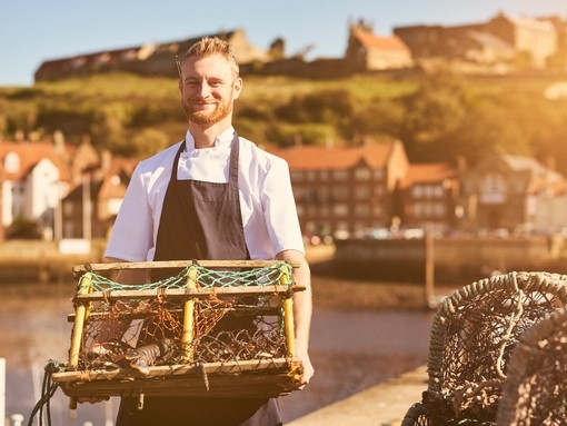 Chef wearing apron on pier holding lobster trap with lobster