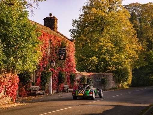 A classic car drives past a pub covered with ivy leaves