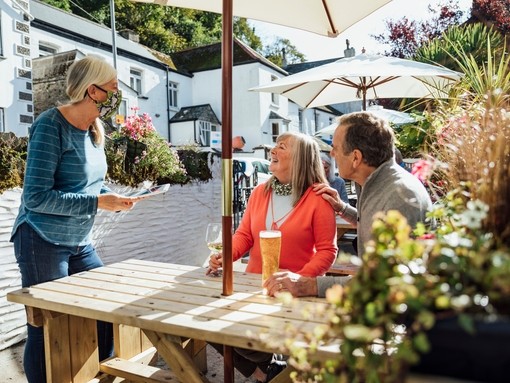 A couple sitting at a wooden table in a beer garden, while a waitress wearing a Covid mask takes their order