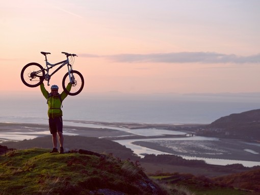 Man holding his mountain bike above his head in celebration at the top of a hill with the coast and sea below him