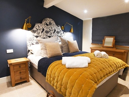 A double bedroom at Rowley Farm Holidays, Worcestershire. The bedroom walls are dark blue and the bed has a mustard yellow padded beadspread. On over-night bag is positioned at the corner of the bed in the foreground. Bronze winner of the New Tourism Business of the Year in the VisitEngland Awards for Excellence 2022