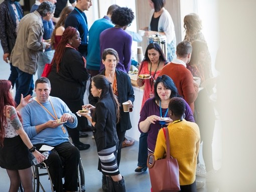 A diverse group of people is networking during a conference break while drinking coffee and eating sandwiches and cake. The focus of the image is on a group of three, two woman standing and one man sitting in a wheelchair, talking to each other in front of people standing at a buffet in the background.