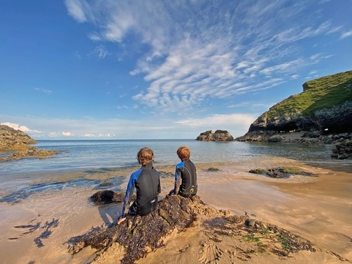 Back view of two young boys in wetsuits sitting on a rock looking out to sea with blue sky