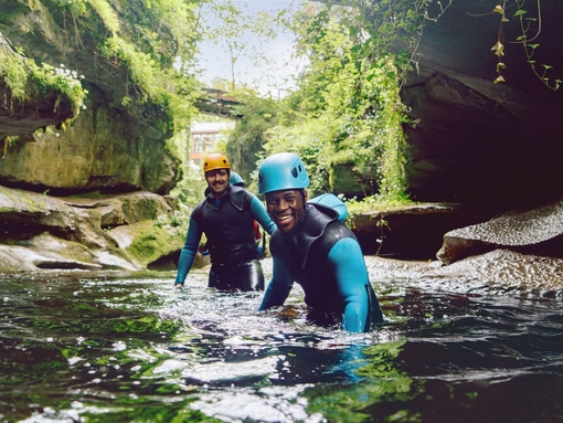 Two men wearing wetsuits and helmets wade waist deep in a gorge