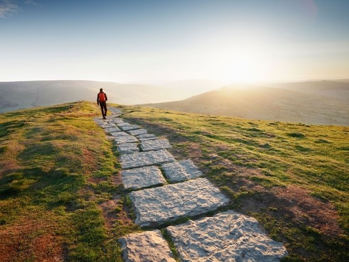 Man in red jacket walking away into the sun at the top of a mountain path
